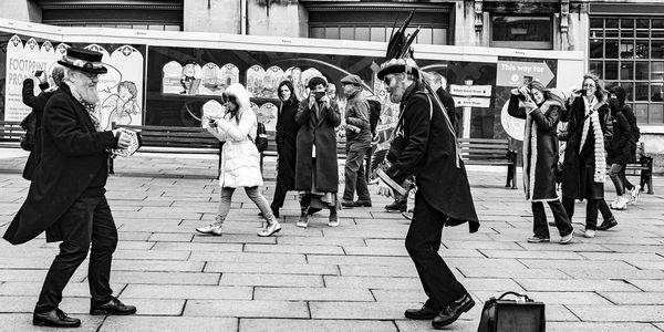 Two men in dark suits and top hats adorned with feathers, play accordion in the street with audience