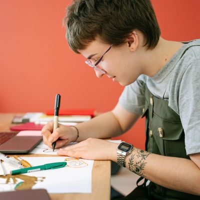 A younger person sits at a table in a bright room surrounded by paper and pens