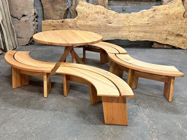 Round table with curved benches to match sitting inside a wood shop with live edge slabs behind it.