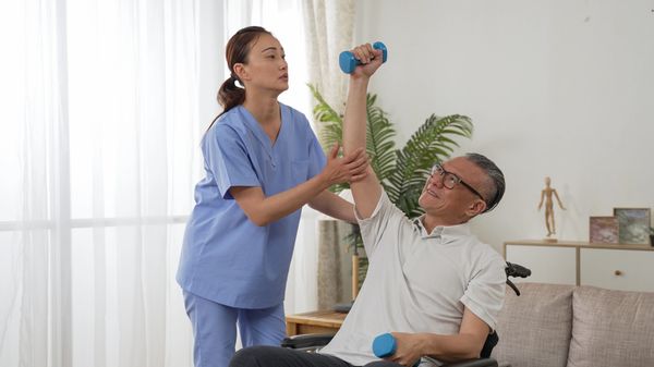 Physical therapist assists an elderly man in a wheelchair with arm exercises using dumbbells.