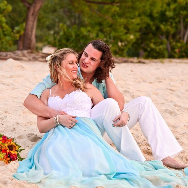 Bride sat in front of groom on a sandy beach wearing a white lace and aqua chiffon wedding dress