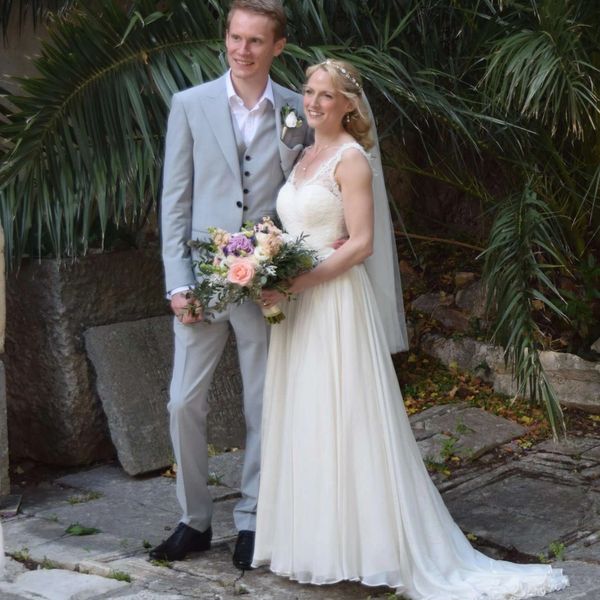 Bride in silk and lace wedding dress with groom in grey suit standing in front of palm trees