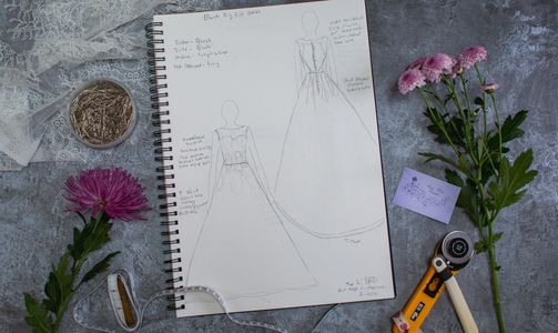 Pencil drawing of wedding dress in a sketchbook surrounded by sewing equipment and pink flowers