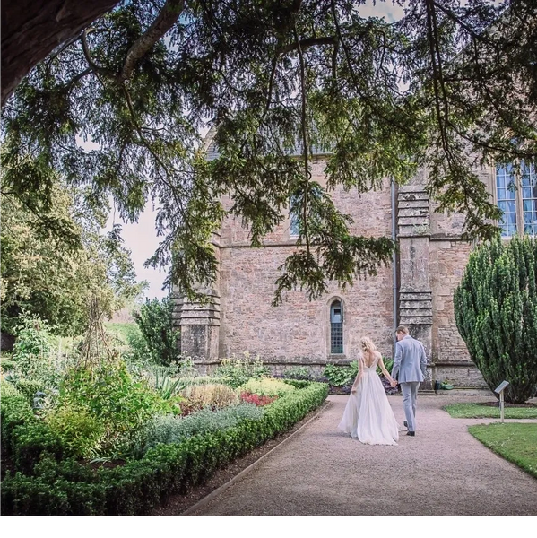 Bride in silk and lace wedding dress and groom walking away along a path in formal gardens