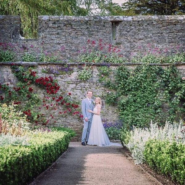 Bride in silk and lace wedding dress standing with groom in front of a wall in a formal garden