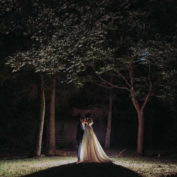 Bride in caped wedding dress and groom in silhouette at nighttime surrounded by trees.