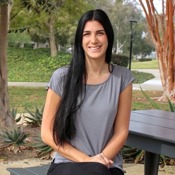 Young woman with long black hair sitting on a bench in a park, smiling at the camera.