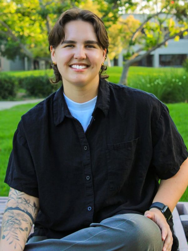 A smiling person with tattoos and short hair sitting outdoors on a bench.
