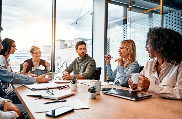 The image captures a diverse group of six professionals engaged in a meeting around a large wooden