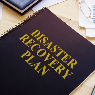 Black spiral notebook titled 'Disaster Recovery Plan' on a desk.