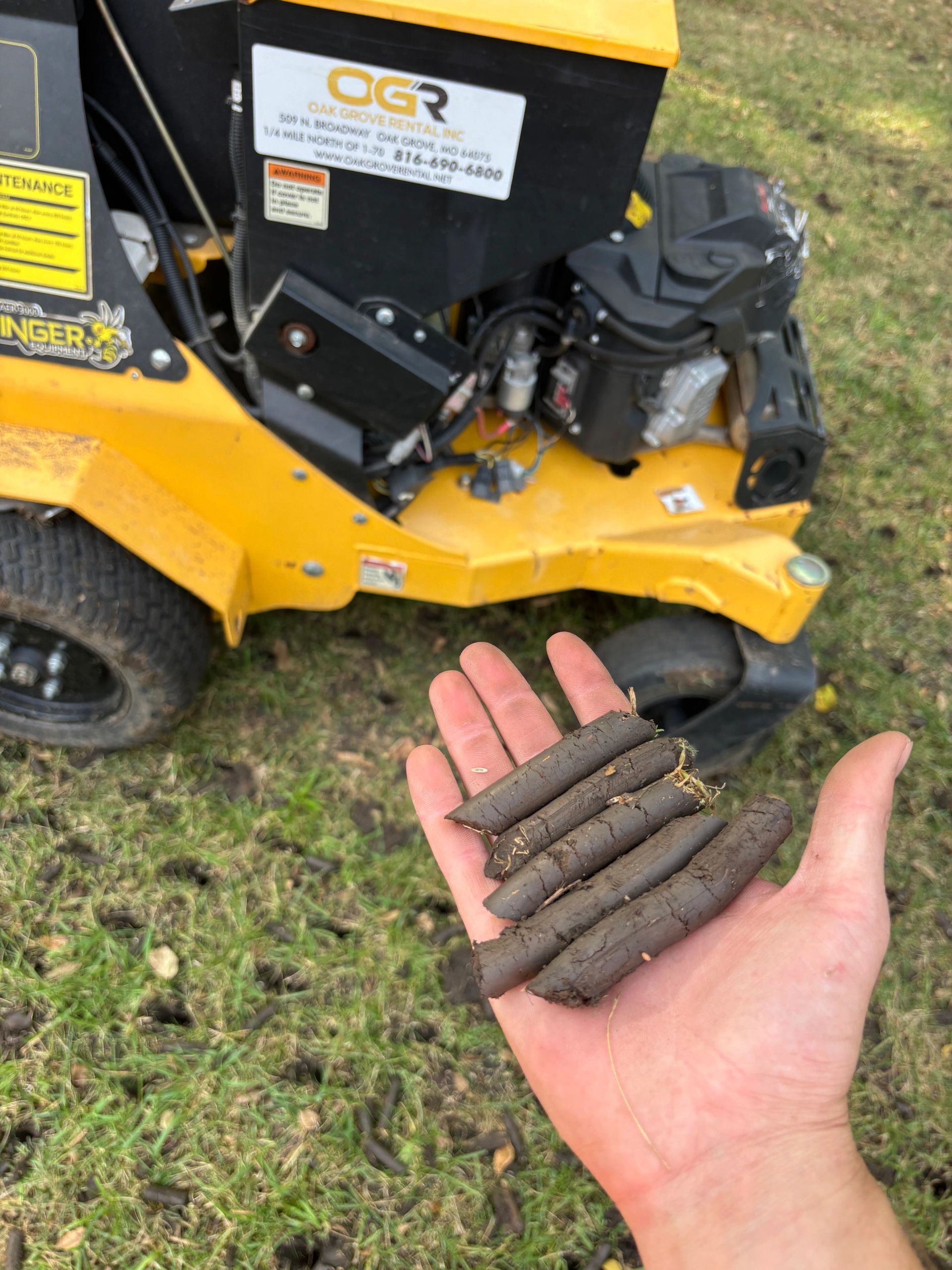 Hand holding cylindrical dirt plugs beside a yellow lawn mower.