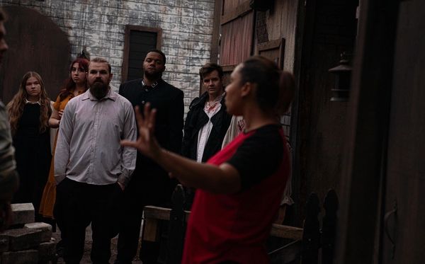 A group listens intently to a woman speaking with her hand raised outdoors.