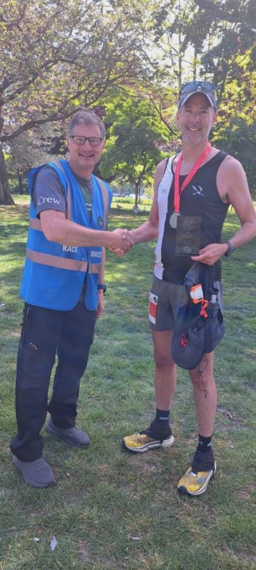 Two men shaking hands, one wearing a race director vest, the other holding a medal and plaque.