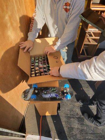 Two people examining a box of Hot Wheels cars beside a skateboard.