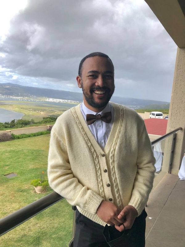 A smiling man in a cream cardigan and brown bow tie stands by a balcony with a scenic view.