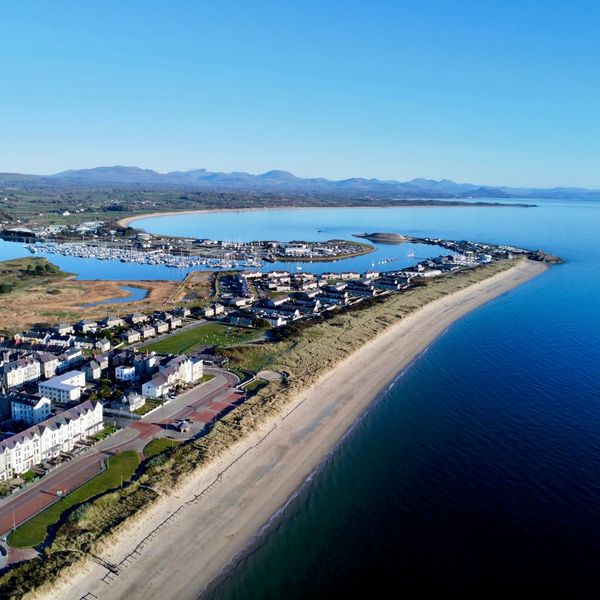A coastal town with a marina, sandy beach, and mountains in the background under a clear blue sky.