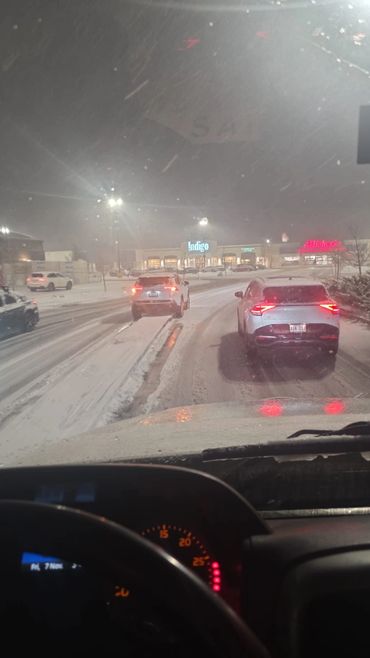 Snowy night with cars driving cautiously in a shopping area parking lot.