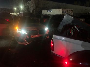 Damaged white BMW with headlights on at night near a pickup truck in a parking lot.