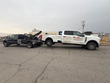 A tow truck towing a white pickup truck with company branding on a cloudy day.