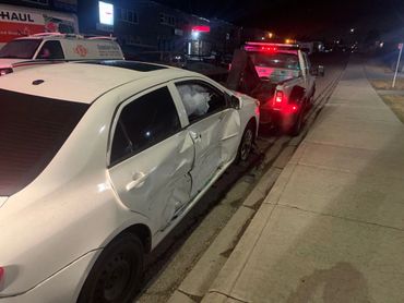White car with side damage being towed at night on a street.
