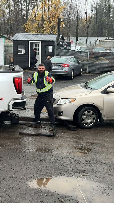 A happy worker in a high-visibility jacket near a car being towed on a rainy day.