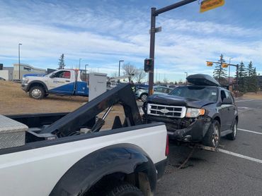 A damaged black SUV being towed by a white tow truck at an intersection.