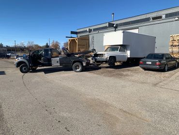 Tow truck towing a damaged white box truck in a parking lot on a sunny day.