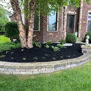 Well-maintained garden bed with a tree, plants, and a Buddha statue in front of a brick house.