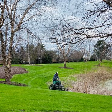 Person mowing grass near a pond on a cloudy day.