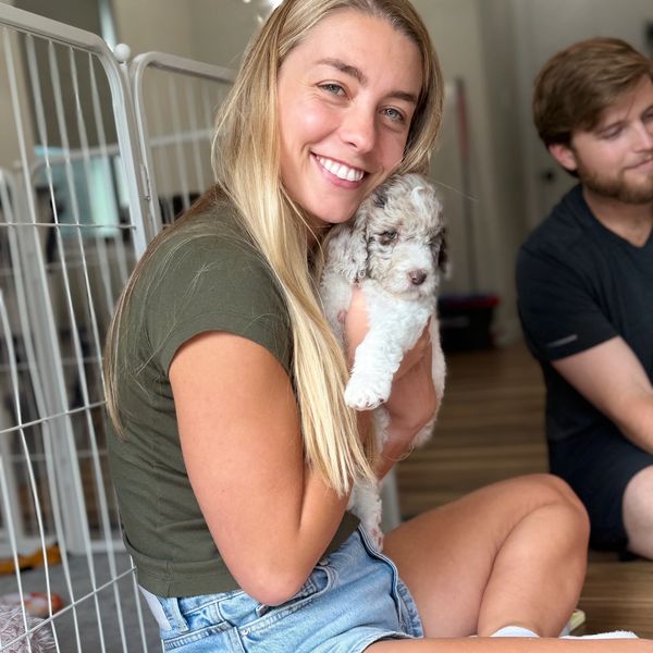 A woman happily holds a small puppy indoors, sitting on the floor.