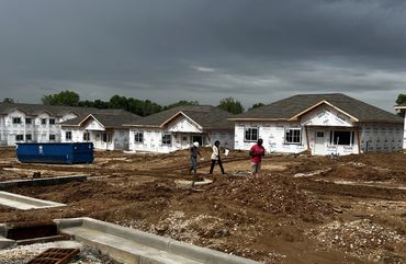 Three people inspect a muddy construction site with partially built houses under cloudy skies.