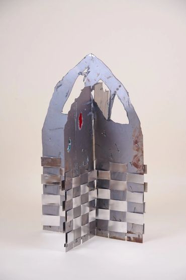 Two planes of metal joined at right angles to stand. At the base, woven metal strips, top is pointed