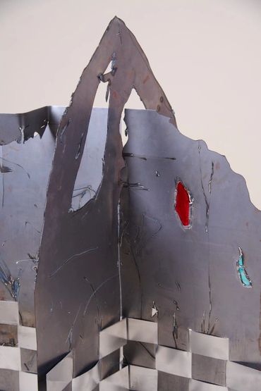 Top section of metal sculpture with rough holes. Two filled with red and blue stained glass