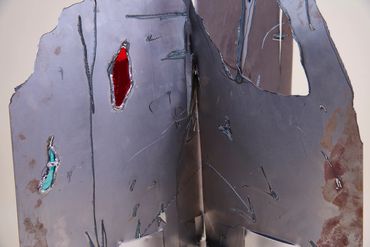 Top section of metal sculpture with rough holes. Two filled with red and blue stained glass