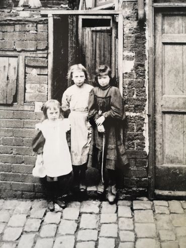 Three children in old B&W photo, wearing long skirts & aprons, stand in doorway of derelict house