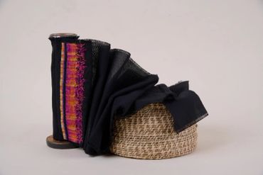 Black cloth, brightly cross-stitched at one end, flows from upright bobbin into handwoven basket