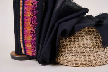 Black cloth, brightly cross-stitched at one end, flows from upright bobbin into handwoven basket