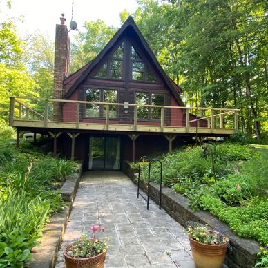 A-frame cabin with large windows surrounded by lush greenery and a stone pathway.