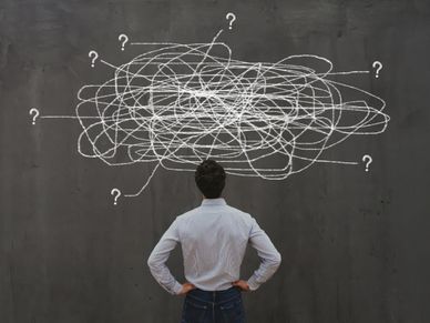 Man standing confused in front of a chaotic web of question marks drawn on a wall.