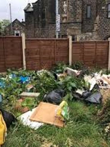 Overgrown garden cluttered with bags and debris against a wooden fence.