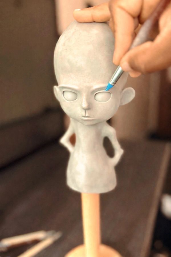 Sculptor adding blue paint to a stylized doll's eye on a clay figure.