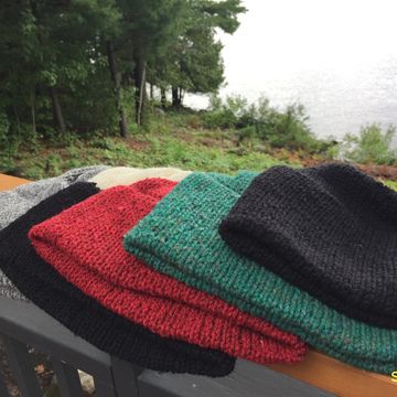 Hand-knit wool hats stacked on an outdoor wooden railing by a lakeside.