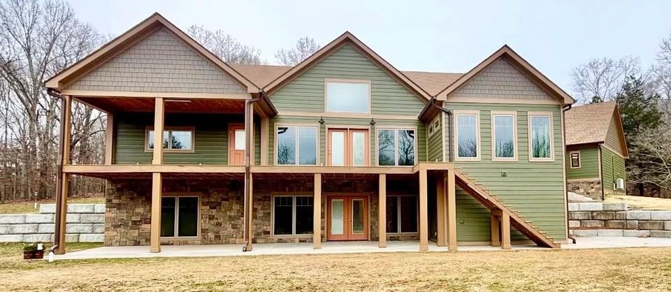 Modern two-story house with green siding and stone accents, surrounded by trees.