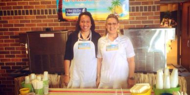 Two women wearing aprons standing behind a juice bar with cups and straws.