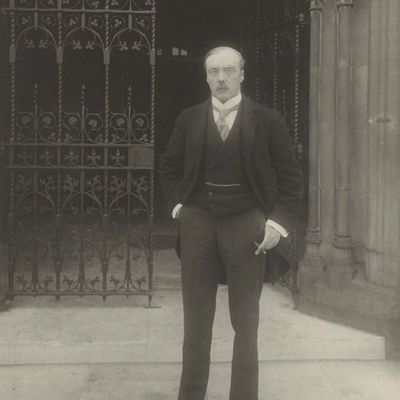 A serious man in formal early 20th-century attire stands outside a gated entrance.