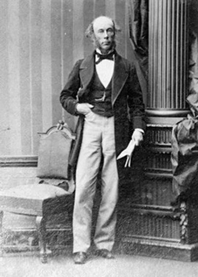 19th-century man in formal attire standing beside a chair and column.