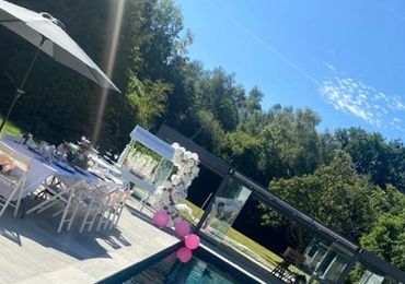 Outdoor poolside party setup with pink balloons and white chairs under a clear sky.