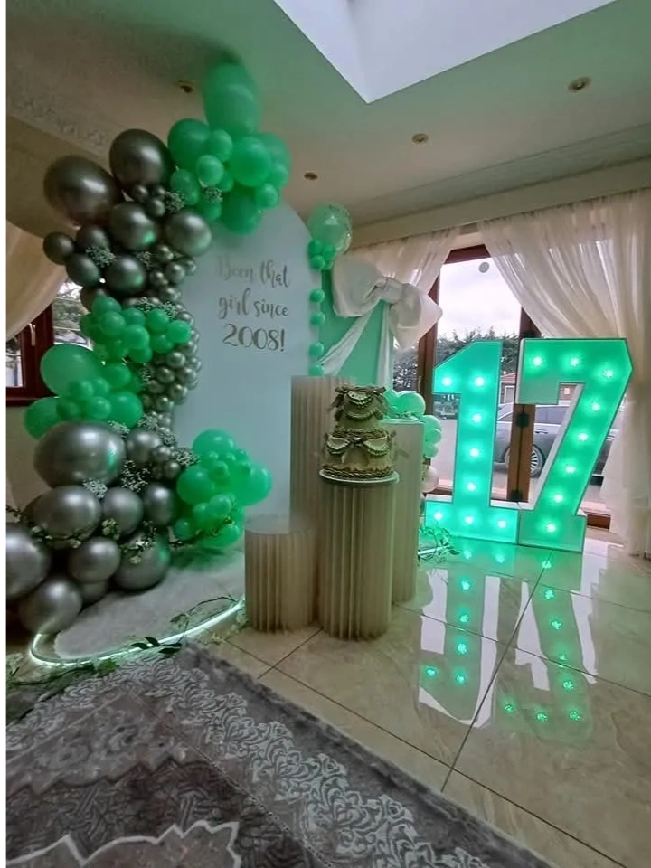Elegant 17th birthday setup with green and silver balloons, cake, and illuminated number 17.