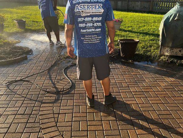 Two workers power washing a brick patio in a backyard.