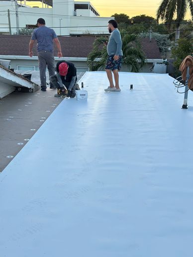 Workers installing a white roofing membrane on a flat roof at sunset.